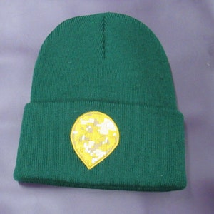 Peut inclure: Un bonnet en tricot vert foncé avec un bord replié. Il présente un écusson jaune avec un motif camouflage dans les tons de beige, blanc et gris. Le bonnet est en tricot côtelé.