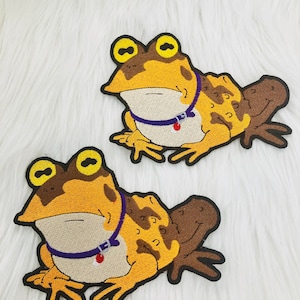 May include: Two embroidered iron-on patches of a cartoon frog wearing a purple collar with a red bell. The frog is brown, yellow, and orange with large yellow eyes.