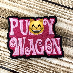 May include: Embroidered patch with the words "Pussy Wagon" in pink, outlined in black. A yellow cat emoji with a smiling face is centered above the word "Wagon". The patch is on a wooden surface.