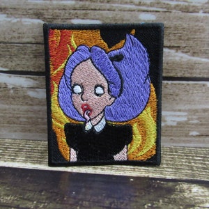 May include: Embroidered iron-on patch featuring a cartoon girl with purple hair and a black dress, set against a background of yellow and orange flames.