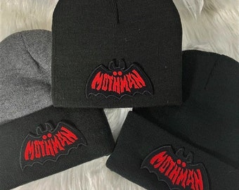 May include: Three black knitted beanies with a red bat graphic and the text "Mothman" on the front. The beanies are on a white fluffy surface.