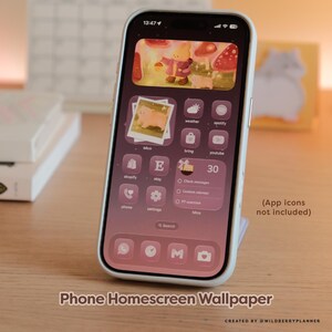 May include: A smartphone with a pink and purple homescreen wallpaper featuring a cartoon bear, mushrooms, and app icons. The phone is in a white case and is propped up on a stand. Text on the screen reads "Phone Homescreen Wallpaper".
