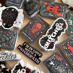 May include: A collection of stickers with gothic and dark themes. Designs include skulls, floral patterns, and text such as "Voracious," "Insatiable," and "I'm in love with a psychopath." The stickers are in various shapes and sizes, with a mix of black, red, and white colors.