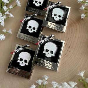 May include: Five silver enamel pins with a black background and a white glitter skull with a red knife. The text on the pins reads "Morally Pitch Black".