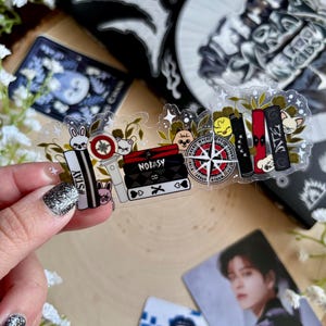 May include: A set of clear stickers featuring various designs, including a compass, a book, a purse, a rabbit, a cat, and a heart with a cross. The stickers have black and white outlines and colorful details. The stickers are arranged in a row on a wooden surface.