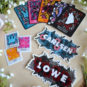 May include: A set of floral stickers with different names, including "Stanford", "NASA", "MIT", "Carlsen", and "Lowe". The stickers are on a wooden surface with white flowers.