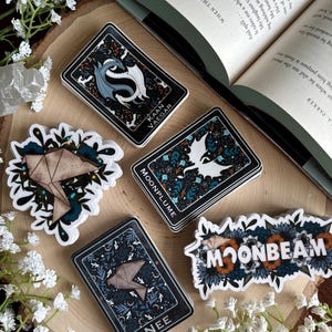 May include: A collection of stickers and tarot cards with celestial and botanical designs. The stickers feature the words "MOONBEAM" and "MOONPLUME". The tarot cards have the words "KAAN VAEGAR" and "NEE". The items are arranged on a wooden surface.