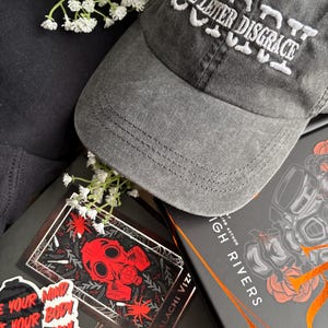 May include: A gray baseball cap with the text "THE LETTER DISGRACE" embroidered in white. The cap is placed on top of a book with a red gas mask graphic and the author's name, Malachi Viz. Decorative stickers and white flowers are also present.