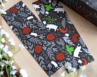 May include: Two black and white bookmark designs with a repeating pattern of red roses, white mice, green herbs, and white pigs. The bookmarks are on a wooden surface.