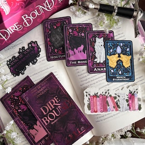 May include: A selection of fantasy-themed items, including a book, cards, and stickers. The book and cards feature the title "Dire Bound" and wolf illustrations. The colour scheme is primarily purple, pink, and white, with floral accents.