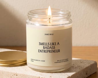 May include: A clear glass jar candle with a lit wick and a cream-colored label. The label features the text JAMES WAX, SMELLS LIKE A BADASS ENTREPRENEUR, SCENT 03 WHITE TEA + JASMINE, and 100% SOY WAX CANDLE HANDPOURED IN CALIFORNIA. A gold lid sits nearby.