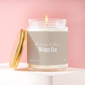 May include: A clear glass candle jar with a lit wick and a gold-colored metal lid placed to the side. The label is light gray with white text that reads Welcome to Your Writer Era, Scent 04, Coconut - Orange, and the code J1185.