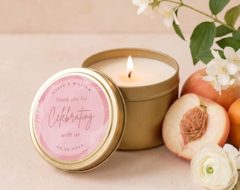 May include: A gold metal candle tin with a lit white candle inside. The lid features a pink circular label with the text "Karen & William", "Thank you for celebrating with us", and "02.02.2022". Nearby are fresh peaches and white flowers.