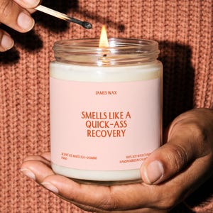 May include: A clear glass jar candle with a light pink label featuring the text JAMES WAX, SMELLS LIKE A QUICK-ASS RECOVERY, SCENT 03, WHITE TEA + JASMINE, F860, and 100% SOY WAX CANDLE HANDPOURED IN CALIFORNIA. The candle has a lit wick.