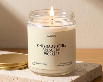 May include: A clear glass jar candle with a lit wick and a cream-colored label. The label reads JAMES WAX, ONLY BAD BITCHES ARE SOCIAL WORKERS, SCENT 03 WHITE TEA + JASMINE, and 100% SOY WAX CANDLE HANDPOURED IN CALIFORNIA. A gold metal lid sits nearby.