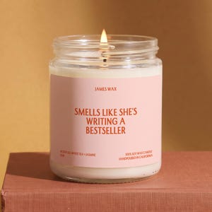 May include: A clear glass jar candle with a light pink label featuring the text JAMES WAX, SMELLS LIKE SHE'S WRITING A BESTSELLER, SCENT 03 WHITE TEA + JASMINE, and 100% SOY WAX CANDLE HANDPOURED IN CALIFORNIA. The candle has white wax and a lit wick.