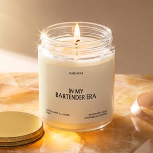 May include: A clear glass jar candle with a lit wick and a cream-colored label. The label features the text JAMES WAX, IN MY BARTENDER ERA, SCENT 03, WHITE TEA + JASMINE, F7268, and 100% SOY WAX CANDLE HANDPOURED IN CALIFORNIA. A gold lid sits nearby.