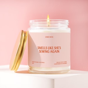 May include: A clear glass jar candle with a lit wick and a gold metal lid resting beside it. The label is light pink with orange text reading JAMES WAX, SMELLS LIKE SHE'S SEWING AGAIN, SCENT 01 ROSE + PATCHOULI, and 100% SOY WAX CANDLE HANDPOURED IN CALIFORNIA.