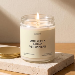May include: A clear glass jar candle with a lit wick and a cream-colored label. The label reads JAMES WAX, SMELLS LIKE A BADASS VETERINARIAN, SCENT 03 WHITE TEA + JASMINE, and 100% SOY WAX CANDLE HANDPOURED IN CALIFORNIA. A gold metal lid sits nearby.