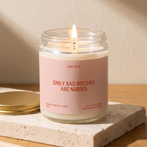May include: A clear glass jar candle with a lit wick and a light pink label. The label features the text JAMES WAX, ONLY BAD BITCHES ARE NURSES, SCENT 03 WHITE TEA + JASMINE, and 100% SOY WAX CANDLE HANDPOURED IN CALIFORNIA in dark red lettering.