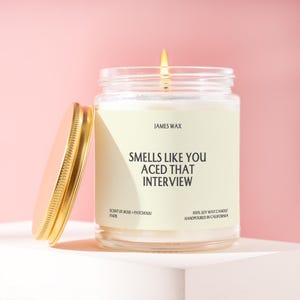 May include: A clear glass jar candle with a gold lid, featuring a cream label that reads JAMES WAX, SMELLS LIKE YOU ACED THAT INTERVIEW, SCENT 01 ROSE + PATCHOULI, and 100% SOY WAX CANDLE HANDPOURED IN CALIFORNIA. The candle has a lit wick.