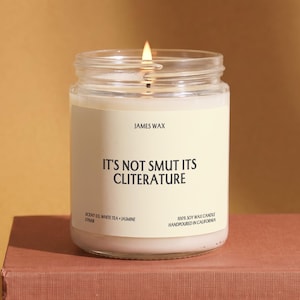 May include: A clear glass jar candle with a white wax fill and a lit wick. The label features the text JAMES WAX, IT'S NOT SMUT ITS CLITERATURE, SCENT 03 WHITE TEA + JASMINE, F7668, and 100% SOY WAX CANDLE HANDPOURED IN CALIFORNIA in black font.