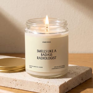 May include: A clear glass jar candle with a lit wick and a cream-colored label. The label features the text JAMES WAX, SMELLS LIKE A BADASS RADIOLOGIST, SCENT 03 WHITE TEA + JASMINE, and 100% SOY WAX CANDLE HANDPOURED IN CALIFORNIA in black font.