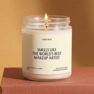 May include: A clear glass jar candle with a white label featuring the text JAMES WAX, SMELLS LIKE THE WORLD'S BEST MAKEUP ARTIST, SCENT 03. WHITE TEA + JASMINE, and 100% SOY WAX CANDLE HANDPOURED IN CALIFORNIA. The candle has a lit wick and white wax.
