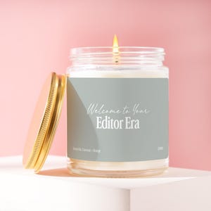 May include: A clear glass candle jar with a lit wick and a gold-colored metal lid placed to the side. The label is sage green with white text that reads Welcome to Your Editor Era, Scent 04, Coconut - Orange, and the code J1101.