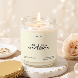 May include: A lit soy wax candle in a clear glass jar with a cream-colored label. The label reads JAMES WAX, SMELLS LIKE A GRANT PROPOSAL, SCENT 03 WHITE TEA + JASMINE, and 100% SOY WAX CANDLE HANDPOURED IN CALIFORNIA. A gold metal lid sits nearby.