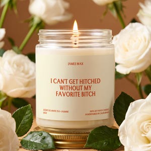 May include: A lit soy wax candle in a clear glass jar with a cream label. The label features the text JAMES WAX, I CAN'T GET HITCHED WITHOUT MY FAVORITE BITCH, SCENT 03. WHITE TEA + JASMINE, J034, and 100% SOY WAX CANDLE HANDPOURED IN CALIFORNIA.
