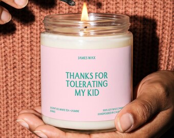 May include: A clear glass jar candle with a light pink label featuring the teal text "JAMES WAX" and "THANKS FOR TOLERATING MY KID". The label also includes "SCENT 03. WHITE TEA + JASMINE" and "100% SOY WAX CANDLE HANDPOURED IN CALIFORNIA".