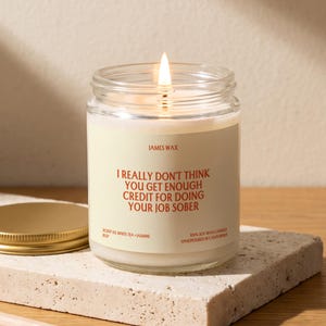 May include: A clear glass jar candle with a lit wick and a cream-colored label. The label features the text JAMES WAX, I REALLY DON'T THINK YOU GET ENOUGH CREDIT FOR DOING YOUR JOB SOBER, SCENT 03 WHITE TEA + JASMINE, and 100% SOY WAX CANDLE HANDPOURED IN CALIFORNIA.