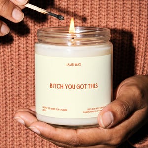 May include: A clear glass jar candle with a cream-colored label featuring the text JAMES WAX, BITCH YOU GOT THIS, SCENT 03, WHITE TEA + JASMINE, J002, and 100% SOY WAX CANDLE HANDPOURED IN CALIFORNIA. The candle has a lit wick and white wax.