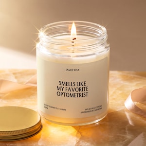 May include: A clear glass jar candle with a white label featuring the text "JAMES WAX" and "SMELLS LIKE MY FAVORITE OPTOMETRIST" along with "SCENT 03, WHITE TEA + JASMINE" and "100% SOY WAX CANDLE HANDPOURED IN CALIFORNIA". A lit wick burns inside.