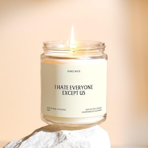 May include: A clear glass jar candle with a lit wick and a cream-colored label. The label features the text JAMES WAX, I HATE EVERYONE EXCEPT US, SCENT 01 ROSE + PATCHOULI, and 100% SOY WAX CANDLE HANDPOURED IN CALIFORNIA in black sans-serif font.