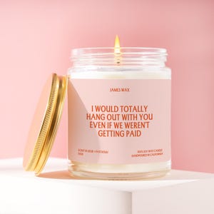 May include: A clear glass jar candle with a lit wick and a gold lid. The label is light pink with dark orange text reading: JAMES WAX, I WOULD TOTALLY HANG OUT WITH YOU EVEN IF WE WEREN'T GETTING PAID, SCENT 01, ROSE + PATCHOULI, F038, 100% SOY WAX CANDLE.