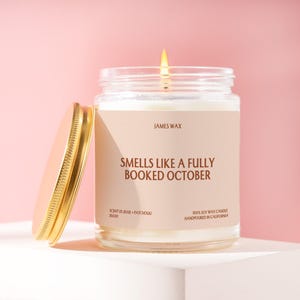 May include: A clear glass jar candle with a gold lid and a beige label. The label reads JAMES WAX, SMELLS LIKE A FULLY BOOKED OCTOBER, SCENT 01 ROSE + PATCHOULI, J6030, and 100% SOY WAX CANDLE HANDPOURED IN CALIFORNIA. A lit wick glows from the center.