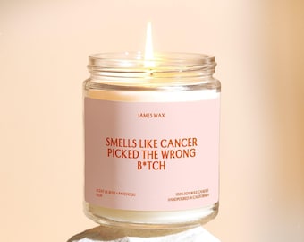 May include: A lit soy wax candle in a clear glass jar with a light pink label. The label features the text JAMES WAX, SMELLS LIKE CANCER PICKED THE WRONG B*TCH, SCENT OF ROSE + PATCHOULI, and 100% SOY WAX CANDLE HANDPOURED IN CALIFORNIA.