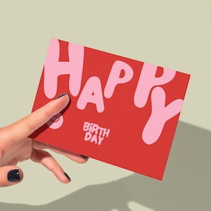 May include: A red birthday card with the word "HAPPY" in large, bubble-style pink letters. Below, the words "BIRTH DAY" are printed in smaller, white letters. The card is held against a light green background.