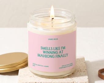 May include: A clear glass jar candle with a lit wick and a pink label. The label features the text JAMES WAX, SMELLS LIKE I'M WINNING AT MAHJONG FINALLY, SCENT 03 WHITE TEA + JASMINE, and 100% SOY WAX CANDLE HANDPOURED IN CALIFORNIA in teal font.