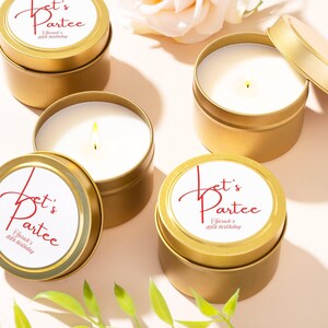 May include: Gold-colored candle tins with white wax and a lit wick. The lids feature a white label with red script that reads "Let's Partee" and "Sarah's 40th birthday". Green leaves and a white flower are in the background.