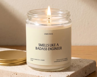 May include: A clear glass jar candle with a lit wick and a cream-colored label. The label reads JAMES WAX, SMELLS LIKE A BADASS ENGINEER, SCENT 03 WHITE TEA + JASMINE, and 100% SOY WAX CANDLE HANDPOURED IN CALIFORNIA. A gold metal lid sits nearby.