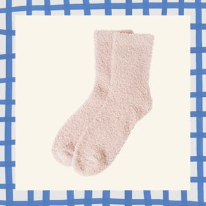 May include: A pair of soft, light pink fuzzy socks. The socks are made of a plush material and are designed for comfort. The socks are a solid color and are displayed on a cream-colored background with a blue and white checkered border.