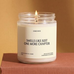 May include: A clear glass jar candle with a lit wick and a cream-colored label. The label reads JAMES WAX, SMELLS LIKE JUST ONE MORE CHAPTER, SCENT 03 WHITE TEA + JASMINE, F1238, and 100% SOY WAX CANDLE HANDPOURED IN CALIFORNIA in black text.
