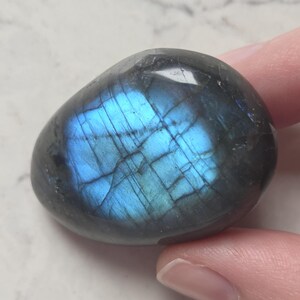 May include: A polished, oval-shaped labradorite gemstone. The stone has a dark gray base color with iridescent blue and teal flashes across its surface. The gemstone is held by a finger.