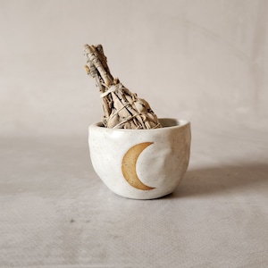 Könnte beinhalten: Weiße Keramikschale mit einem Halbmond-Design in hellbrauner Farbe. Die Schale enthält ein Bündel getrockneten Salbeis.