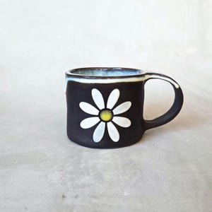 May include: A black ceramic mug featuring a white daisy with a yellow centre. The rim is blue and white. The mug has a curved handle. The mug is suitable for hot beverages.