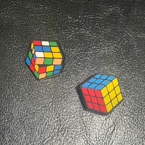 May include: Two colorful Rubik's Cube charms. One is pentagonal, the other is a cube. The cubes feature red, blue, yellow, green, orange, and white squares. The charms have a black border and are on a textured black surface.