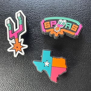 May include: Three decorative shoe charms featuring San Antonio Spurs themes. One charm is a spur logo, one is a text logo reading SPURS, and one is a Texas state map shape divided into teal, orange, and pink sections with a white star.